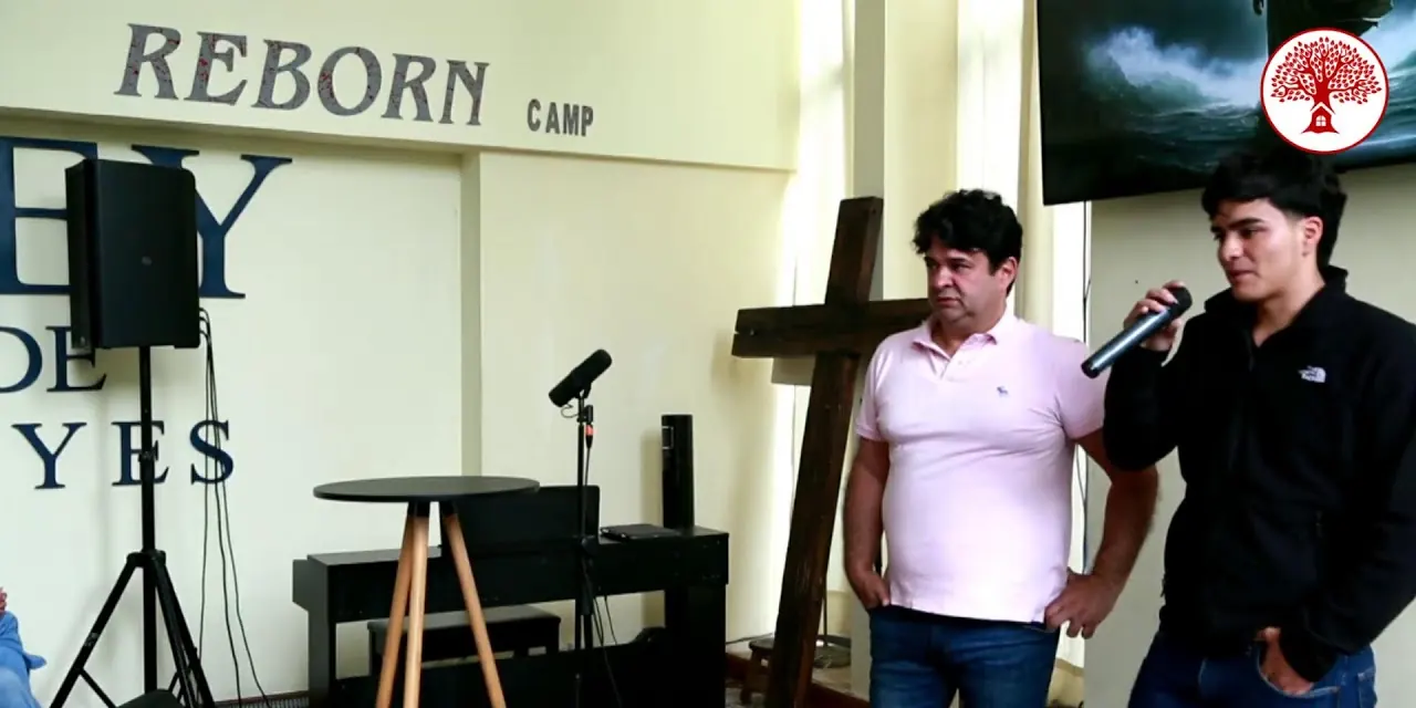 Un joven habla con el micrófono junto a un pastor y a la cruz de madera, bajo el letrero «REBORN CAMP», en el último día de REBORN Camp 2025.